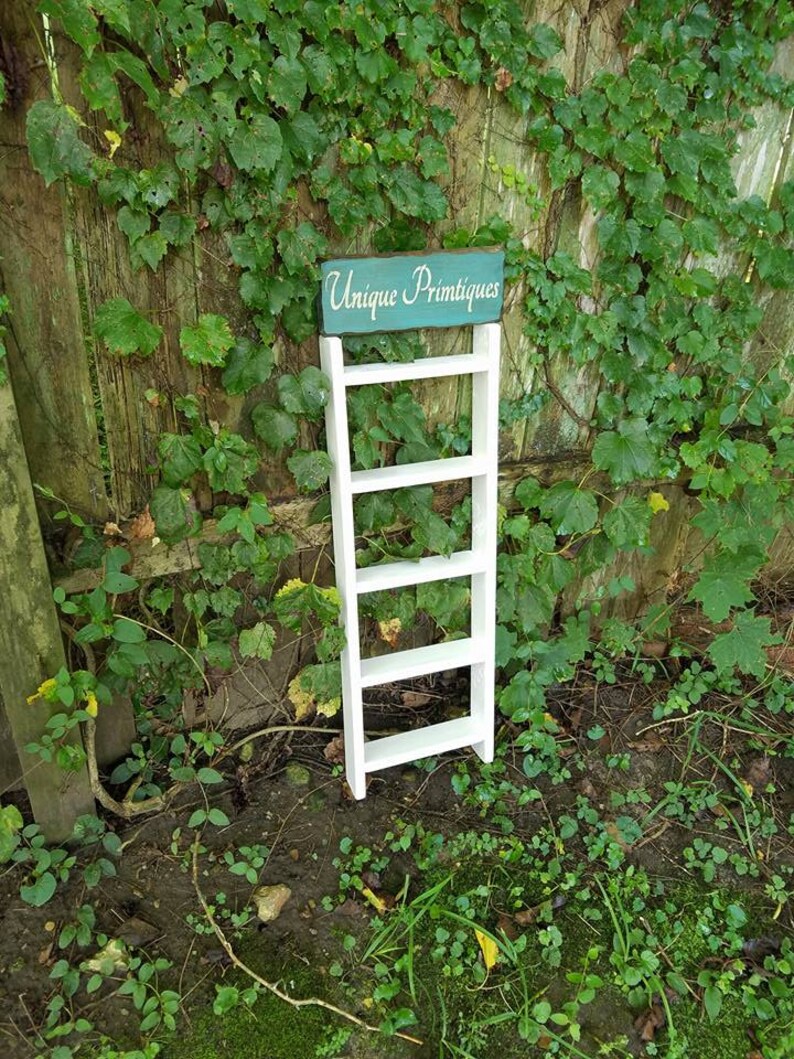 May include: White wooden ladder shelf with five shelves, standing in front of a green ivy covered wooden fence. A teal blue sign with the text "Unique Primitives" is attached to the top of the shelf.