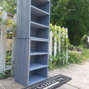 May include: A gray wooden shelf unit with five shelves. The shelf unit is standing on a concrete surface in front of a white picket fence. A black sign with the text "UNIQUE, DISTINCTIVE & RUSTIC FURNITURE SINCE 2018 FARMHOUSE" is lying on the ground in front of the shelf unit.
