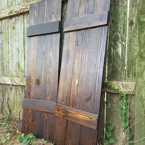 May include: Two dark brown wooden shutters with a rustic finish. Each shutter has three horizontal panels with a raised edge. The shutters are leaning against a wooden fence.