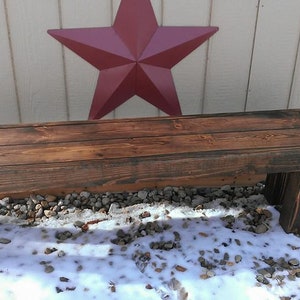 May include: A rustic, stained wooden bench with a rectangular top and four sturdy legs. The bench is set against a white wall with a large red star decoration. The bench is approximately 48 inches long.