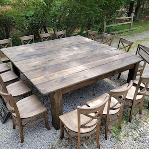May include: A large, rustic wooden table with a square top and a dark brown finish. The table is surrounded by 16 chairs with a simple, cross-back design. The chairs are made of light brown wood and have a distressed finish.