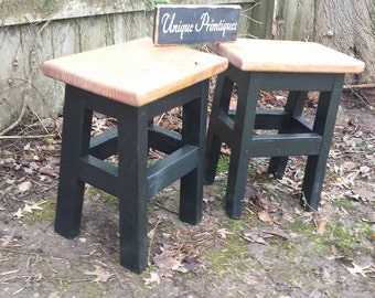 May include: Two black wooden stools with natural wood tops. The stools have a simple, rustic design and are perfect for adding a touch of farmhouse style to any room.