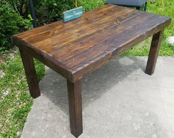 May include: A rectangular wooden table with a dark brown stained finish and a visible wood grain pattern. The table features four sturdy square legs and a flat top surface. A small rectangular sign with the text Unique Primtiques is placed on top.