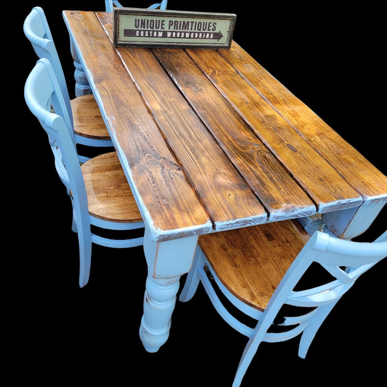 May include: A rustic wooden dining table with four matching chairs. The table has a distressed brown tabletop and white-painted legs. The chairs are light blue with wooden seats. A sign above the table reads "UNIQUE PRIMTIQUES CUSTOM WOODWORKING".