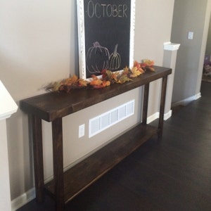 May include: A dark brown wooden console table with a single shelf. The table is decorated with a fall-themed garland of orange and brown leaves. A chalkboard sign with the word "OCTOBER" written on it is hanging on the wall behind the table.