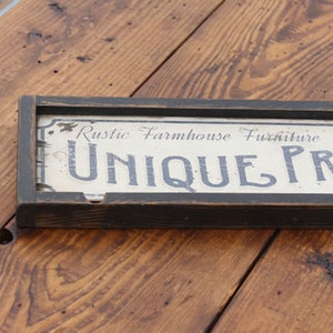 May include: A rectangular wooden sign with a black frame, featuring the text "Rustic Farmhouse Furniture UNIQUE PRINTS" in a distressed, vintage style. The sign is set against a wooden surface.