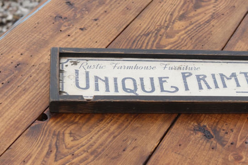 May include: A rectangular wooden sign with a black frame, featuring the text "Rustic Farmhouse Furniture UNIQUE PRINTS" in a distressed, vintage style. The sign is set against a wooden surface.