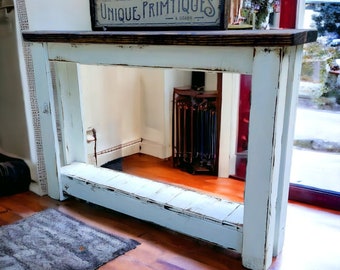 May include: A white painted console table with a distressed finish and a dark brown top. The table has a shelf and is sitting on a wooden floor. A sign that reads "Unique Primitives" is on top of the table.