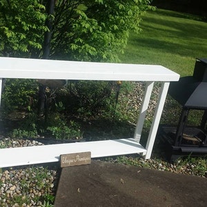 May include: White console table with a single shelf and a small wooden sign that reads "Unique Boutique". The table is sitting on a gravel surface next to a black metal fire pit.