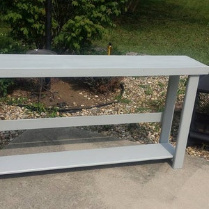 May include: A gray wooden console table with two shelves. The table is in a backyard setting with gravel and green grass.