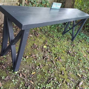 May include: Black wooden console table with a black X-shaped base. The table is sitting on a bed of green moss and leaves.