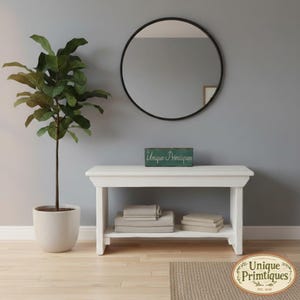 May include: A white wooden console table with a lower shelf holding stacks of folded light-colored linens. A green rectangular sign with the text Unique Primtiques sits on the tabletop. A round black-framed mirror hangs on the wall above.