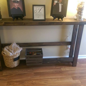 May include: A dark brown wooden console table with a shelf. The table has a distressed finish and is decorated with framed pictures and a vase with flowers. The table is sitting on a hardwood floor.
