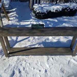 May include: A dark stained wood console table with a single shelf. The table has a rustic farmhouse style and is sitting on a snowy surface. The text "Unique  Antiques" is visible on a small sign in front of the table.