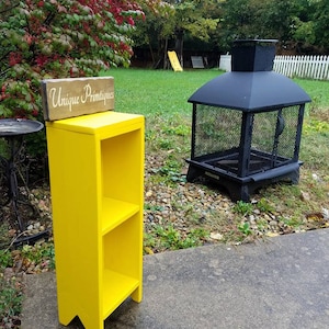 May include: A yellow wooden shelf with two shelves and a sign that says "Unique Primitives".