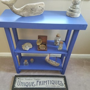 May include: A blue wooden shelf unit with three tiers, displaying nautical-themed decor. Items include a whale sculpture, a lighthouse, seashells in jars, a small sailboat, and a sign that reads "UNIQUE PRIMITIQUES & DECOR."