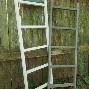 May include: Two wooden ladders, one painted white and the other gray, leaning against a wooden fence. The white ladder has a sign that reads "Unique Primitives".