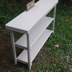 May include: White wooden shelf unit with two shelves. The shelf unit is painted white and has a rustic look. The shelf unit is sitting on a bed of green grass. A small wooden sign with the text "Unique Treasures" is sitting on top of the shelf unit.