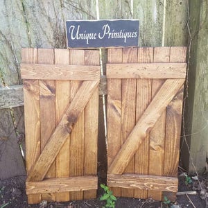 May include: Two wooden barn doors with a distressed brown finish. The doors are made of individual planks of wood and have a diagonal brace across the center. The doors are closed and have a sign above them that reads "Unique Primitives".