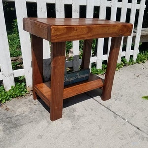 May include: A brown wooden side table with a rectangular top and a lower shelf. The table features four thick, straight legs and a smooth, stained finish. A small, rectangular blue sign with the white text Unique Primtiques sits on the lower shelf.