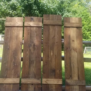 May include: Four wooden shutters in a rustic brown color, leaning against a wooden fence. The shutters are each made of four horizontal planks with a single horizontal plank across the top. The shutters are in front of a green grassy area and a wooden fence. A black sign with white text reads "Unique Distinctive Farmhouse" in front of the shutters.
