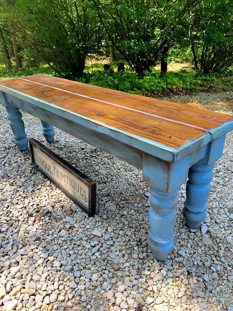 May include: A rustic wooden bench with a distressed blue painted base and legs. The bench top is stained a warm brown with a light-colored stripe down the center. A sign that reads "UNIQUE PRIMTIQUES" is leaning against the bench.