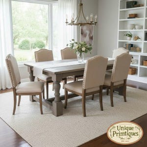 May include: A rectangular wooden dining table with a light gray finish and thick, turned legs, accompanied by six upholstered chairs in a neutral beige fabric with nailhead trim, arranged around the table in a bright, open dining room setting.