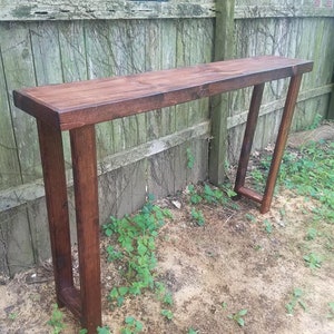 May include: A dark brown wooden console table with a long, rectangular top and two vertical supports. The table is standing on a dirt and grass surface in front of a wooden fence.