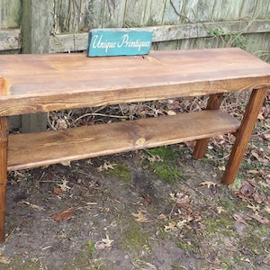May include: A rustic wooden console table with a shelf, stained in a dark brown color. The table has a simple design with straight legs and a rectangular top. The table is sitting on a bed of brown leaves and green grass.