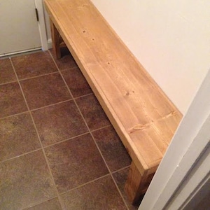 May include: A light-colored wooden bench with a rectangular top and four sturdy legs. The bench is made of unfinished wood and is placed against a white wall. The bench is approximately 60 inches long.