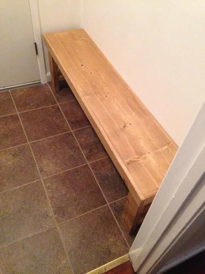 May include: A light-colored wooden bench with a rectangular top and four sturdy legs. The bench is made of unfinished wood and is placed against a white wall. The bench is approximately 60 inches long.