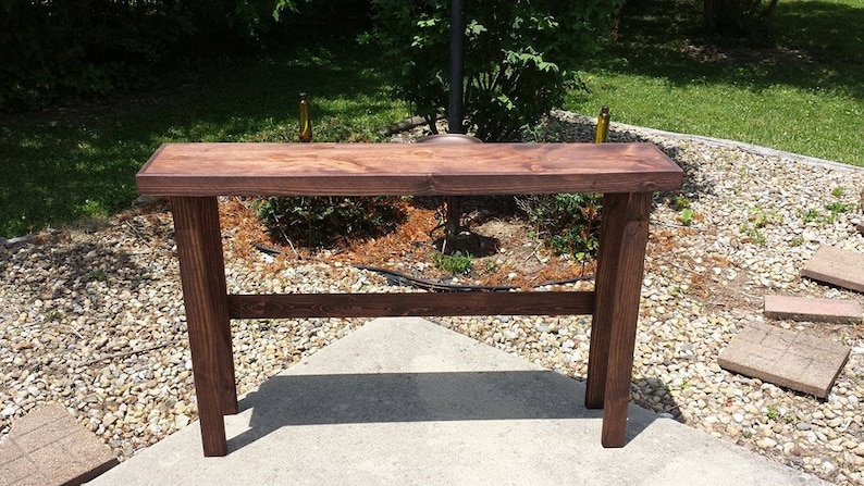 K&ouml;nnte beinhalten: Ein dunkelbrauner Holz-Konsolentisch mit einem einzigen Regal. Der Tisch steht auf einer Kiesfl&auml;che.