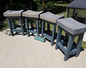 May include: Four gray wooden stools with a curved seat and a dark blue paint finish. The stools are arranged in a row on a concrete surface. The sign "Unique Pentiques" is visible in the foreground.