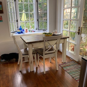 May include: A rectangular dining table with a dark brown wood top and a distressed white painted base, accompanied by two matching chairs featuring an X-back design. The table is positioned in a bright room with large windows and a glass door.