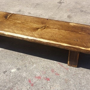 May include: A small, rectangular wooden bench with a dark brown stain. The bench is supported by two short legs and measures approximately 24 inches long. The edges have a lighter, natural wood color.