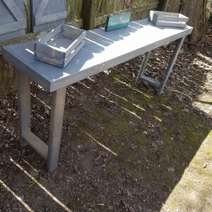 May include: A gray wooden console table with a distressed finish. The table has a long rectangular top and two legs on each side. The table is sitting on a grassy surface with a wooden sign that says "Unique Boutique" on top of the table.