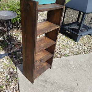 May include: A dark brown wooden shelf unit with four shelves. The shelf unit is standing on a concrete surface. A teal sign with white lettering reads "Unique Primtiques".