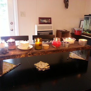 May include: A wooden shelf with six candles, including a red jar candle, a clear glass jar candle, a brown glass jar candle, a clear glass bottle candle, and two metal tin candles. The shelf is sitting on a black table with two yellow and green patterned coasters.