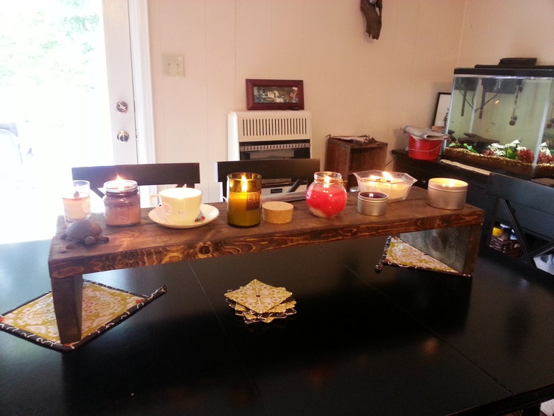 May include: A wooden shelf with six candles, including a red jar candle, a clear glass jar candle, a brown glass jar candle, a clear glass bottle candle, and two metal tin candles. The shelf is sitting on a black table with two yellow and green patterned coasters.