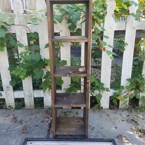 May include: A dark brown wooden shelf unit with four shelves. The shelf unit is standing in front of a white picket fence with green vines growing on it. A sign with the text "Rustic Farmhouse Furniture Unique Primitiques & D&eacute;cor" is lying on the ground in front of the shelf unit.