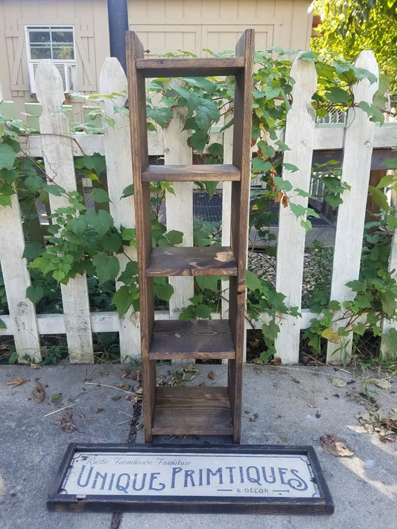 May include: A dark brown wooden shelf unit with four shelves. The shelf unit is standing in front of a white picket fence with green vines growing on it. A sign with the text "Rustic Farmhouse Furniture Unique Primitiques & D&eacute;cor" is lying on the ground in front of the shelf unit.