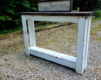 May include: White console table with a distressed finish and a dark brown top. The table has a shelf and is sitting on a gravel surface. A sign that reads "Unique Primitives" is sitting on top of the table.