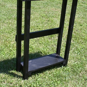 May include: A black wooden console table with a simple design. It has a rectangular top and a shelf underneath. The table is standing on a grassy surface.