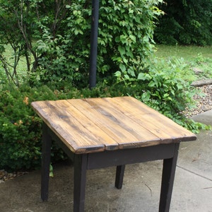 May include: A rustic wooden table with a dark brown stain and a light wood top. The table has four legs and is sitting on a concrete patio.