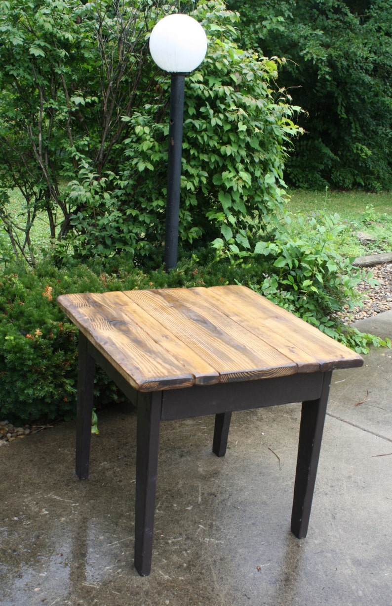 May include: A rustic wooden table with a dark brown stain and a light wood top. The table has four legs and is sitting on a concrete patio.