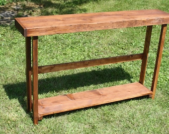 May include: A rustic wooden console table with a natural wood finish. The table has a rectangular top and two shelves. The table is made of solid wood and has a simple, elegant design.