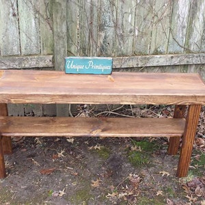 May include: A rustic wooden bench with a shelf, stained in a dark brown color. The bench is sitting on a bed of brown leaves and dirt. A small sign with the text "Unique Primtiques" is sitting on top of the bench.