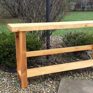 May include: A rustic wooden console table with two shelves. The table is made of natural wood and has a light brown finish. It is a simple and functional piece of furniture that would be perfect for a hallway or entryway.