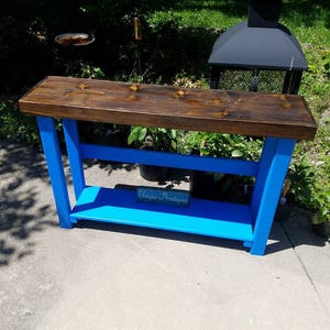 May include: A blue console table with a wooden top and a lower shelf. The table is painted a bright blue and has a rustic wood top. The table has a sign that says "Unique Primtiques" on the shelf.
