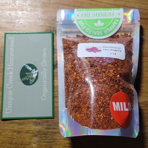 May include: A small resealable pouch of Gochugaru Chili Powder, labeled "MILD" and "1 oz", with a red and white label that says "Gochugaru Chili Powder 1 oz" and a green label that says "TAMPER SEALED FOR FRESHNESS". The pouch is on a wooden surface.